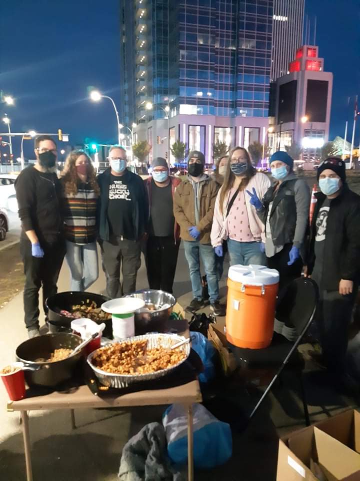 group photo of 9 individuals standing behind serving table with food on it. It is night time and they are standing in downtown edmonton, across the street from Boyle Street community services and in front of some sky scrapers.