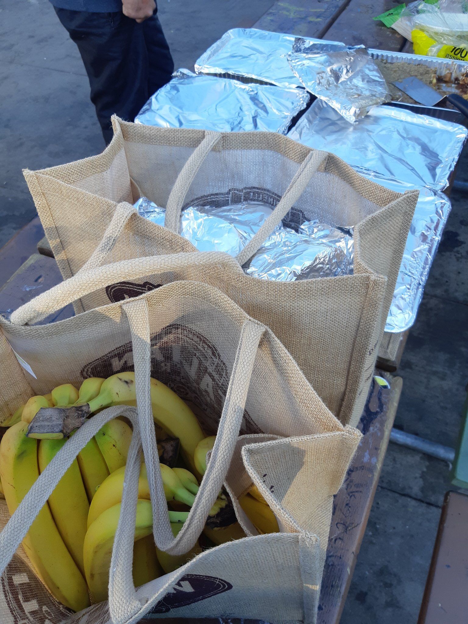 An image of the food to be served - canvas bags, one filled with bananas and another filled with foil-wrapped food in the foreground. Large foil dishes wrapped with food a bit further away from the camera. Paper plates and serving utensils. All piled on top of a picnic table.